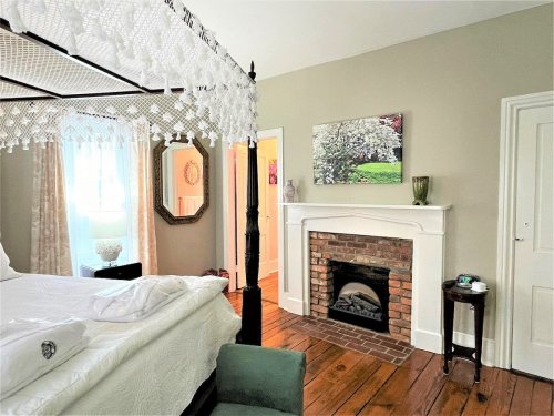 Cozy bedroom with a four-poster bed, fireplace, and light-colored walls, featuring wooden floors and soft natural light.