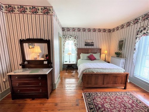 Cozy bedroom with floral wallpaper, a wooden bed, dresser, and natural light from windows.