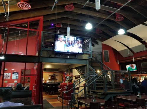 Interior of a restaurant with exposed beams, a large TV showing news, and a staircase leading to an upper level.