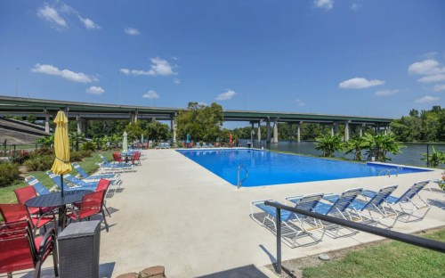 A sunny pool area with lounge chairs, umbrellas, and a view of a river and bridge in the background.