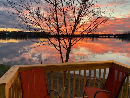 A serene lakeside view at sunset, featuring a tree, two chairs, and vibrant reflections on the water.