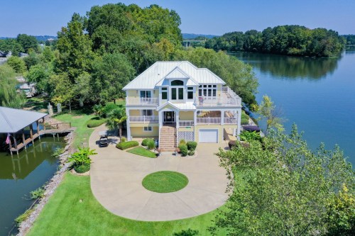 A large waterfront home with a circular driveway, surrounded by trees and a calm lake under a clear blue sky.