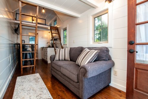 Cozy interior of a tiny house featuring a gray sofa, wooden floors, and a lofted sleeping area with a ladder.