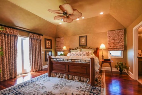 Cozy bedroom with a wooden bed, decorative curtains, and warm lighting, featuring a ceiling fan and a plush rug.
