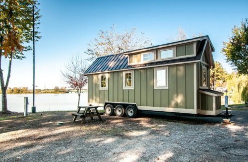 A small green and beige tiny house on wheels parked by a lake, surrounded by trees and a clear blue sky.