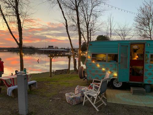 A vintage teal camper by a lake at sunset, surrounded by trees and string lights, with cozy seating nearby.