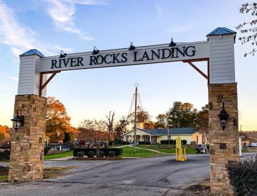 Sign for River Rocks Landing, framed by stone pillars, with trees and a building in the background during sunset.
