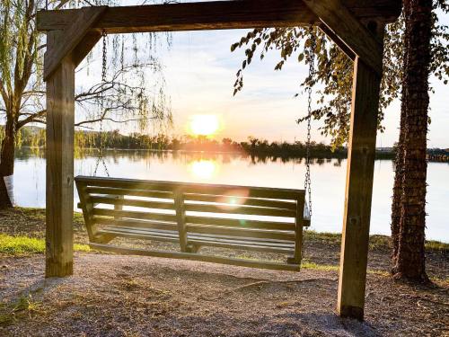 A wooden swing hangs by a lake at sunset, with trees framing the serene scene and sunlight reflecting on the water.