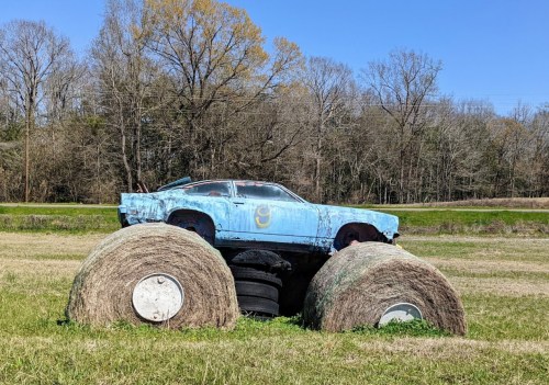 A blue car perched on large hay bales and tires in a grassy field under a clear blue sky.