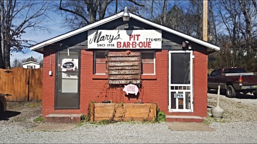 A small brick building with a sign reading "Mary's Pit Bar-B-Que," featuring an open sign and gravel parking area.