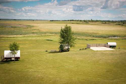 A wide-open landscape featuring green fields, a few trees, and a small wooden deck under a cloudy sky.