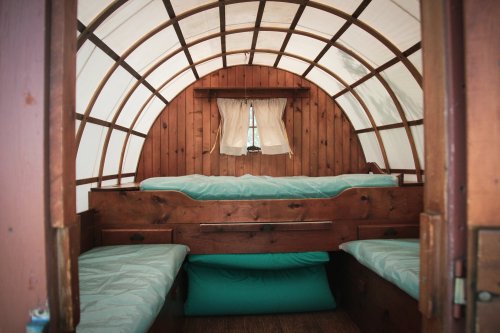 Interior of a cozy wooden wagon with two beds and a window, featuring a curved ceiling and soft bedding.