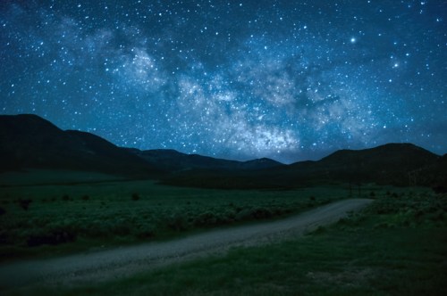 A starry night sky filled with the Milky Way over a dark, mountainous landscape and a gravel road.