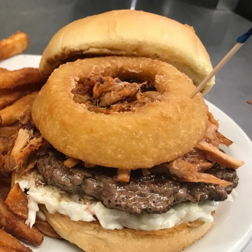 A burger with a beef patty, pulled pork, coleslaw, and a crispy onion ring, served with sweet potato fries.