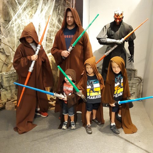 A group of adults and children dressed as Jedi, holding lightsabers, with a Sith figure in the background.