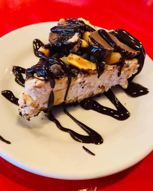 A slice of dessert topped with chocolate drizzle, peanut butter cups, and crushed nuts on a white plate.