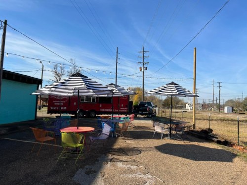A food truck with a red exterior parked in an outdoor area with colorful tables and striped umbrellas under a clear blue sky.