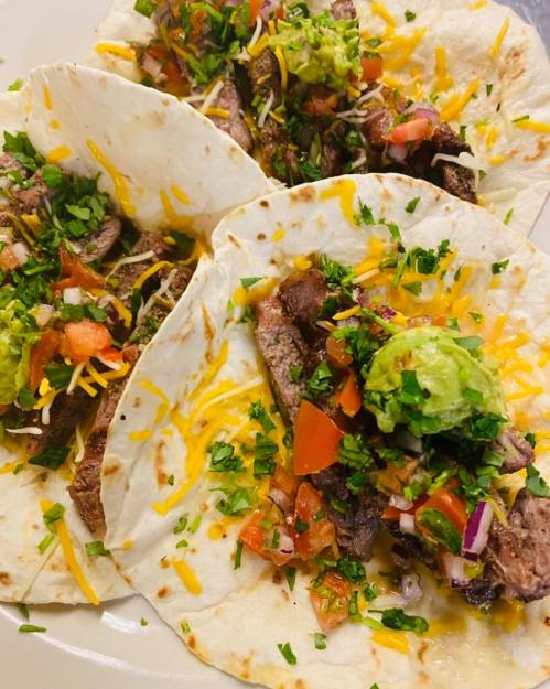 Three tacos filled with grilled meat, cheese, guacamole, pico de gallo, and fresh herbs on soft tortillas.