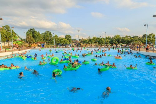 A crowded water park pool filled with people on green floats, surrounded by trees and blue skies.