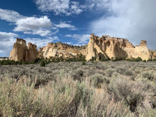A scenic view of rugged cliffs and rock formations against a blue sky, surrounded by sagebrush and sparse vegetation.