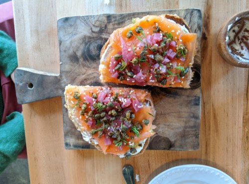 Two slices of toast topped with smoked salmon, diced tomatoes, herbs, and capers on a wooden cutting board.