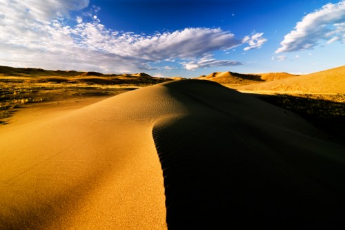 A vast desert landscape with rolling sand dunes under a blue sky and scattered clouds.