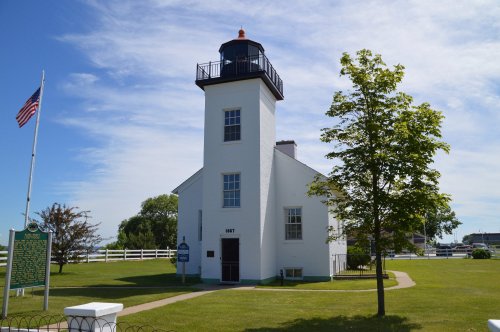 A white lighthouse with a black lantern, surrounded by green grass and trees, under a blue sky with clouds.