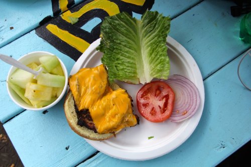 A plate with a cheeseburger topped with two slices of cheddar, lettuce, tomato, onion, and a side of fruit salad.