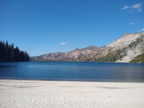 A serene lake surrounded by mountains and pine trees, with a sandy beach in the foreground under a clear blue sky.