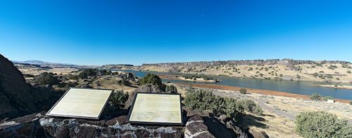 Panoramic view of a river valley with two informational plaques in the foreground under a clear blue sky.