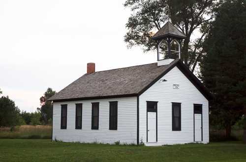 A small, white, historic schoolhouse with a bell tower, surrounded by green grass and trees.