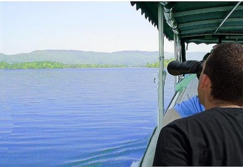 A person on a boat gazes through binoculars at a calm lake surrounded by green hills under a clear sky.