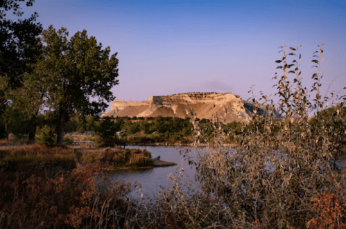 Scenic view of a river with a rocky cliff and trees under a clear blue sky at dusk.