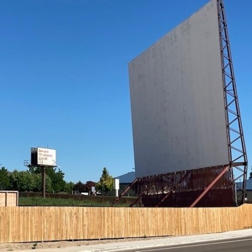 A large blank billboard stands next to a wooden fence, with a smaller sign in the background against a clear blue sky.