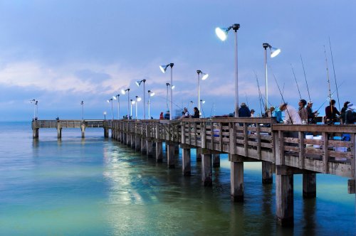 A fishing pier at dusk, illuminated by lights, with people fishing along the wooden structure over calm waters.