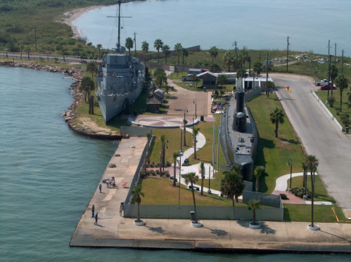 Aerial view of a waterfront park featuring a naval ship and submarine, surrounded by palm trees and pathways.