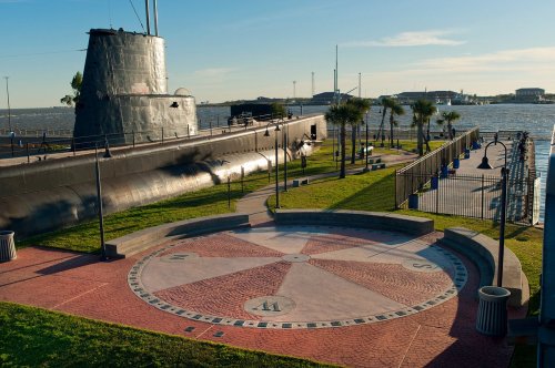 A submarine docked by a waterfront park, featuring a compass rose design on the ground and palm trees nearby.