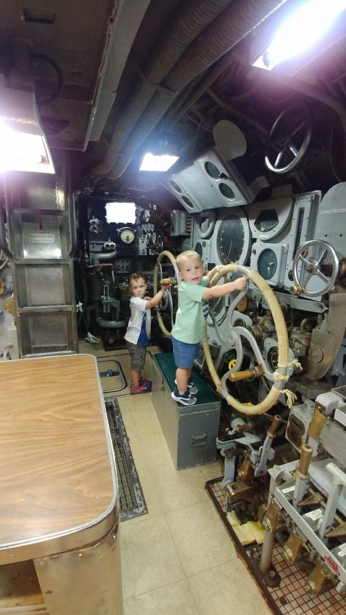 Two young children play with large steering wheels inside a submarine, surrounded by various controls and machinery.