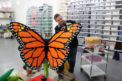A person adjusts a large butterfly sculpture made of colorful building blocks in a workshop filled with storage bins.
