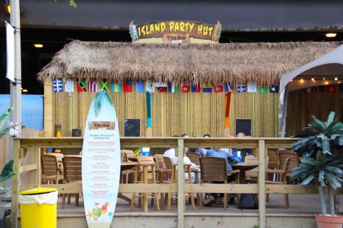 A tiki bar with a thatched roof, flags from various countries, and outdoor seating under a wooden fence.