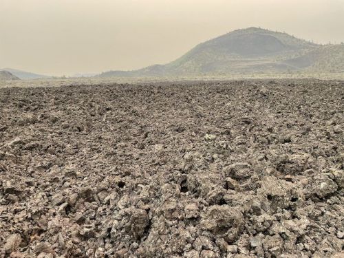 A barren landscape of rough, dark volcanic rock with a distant hill under a hazy sky.