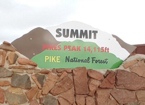 Sign at the summit of Pikes Peak, displaying "Pikes Peak 14,115 ft" and "Pike National Forest" on a rocky background.