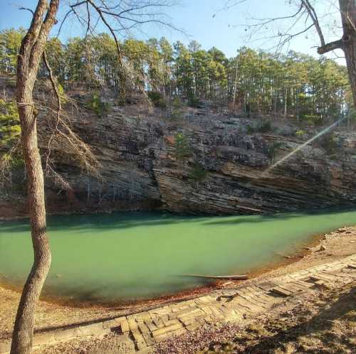 A serene river flows beside a rocky cliff, surrounded by lush green trees under a clear blue sky.