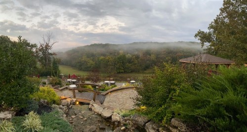 A scenic view of a misty landscape with rolling hills, greenery, and a stone patio in the foreground.