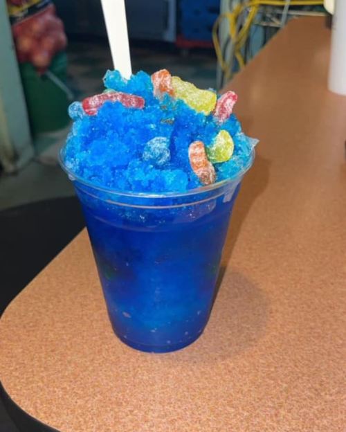 A clear cup filled with blue shaved ice topped with colorful gummy candies on a table.