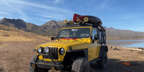 A yellow off-road vehicle parked by a lake, with mountains in the background and a canoe on the roof.