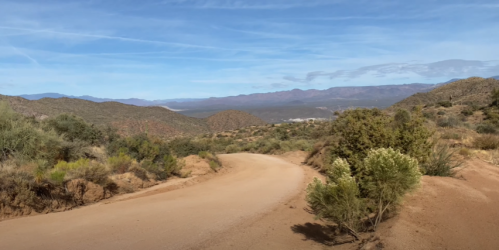 A winding dirt road through a hilly landscape with sparse vegetation and distant mountains under a clear blue sky.