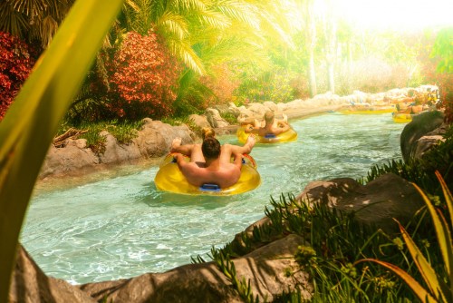 People relax on yellow inner tubes in a winding lazy river surrounded by lush greenery and bright sunlight.