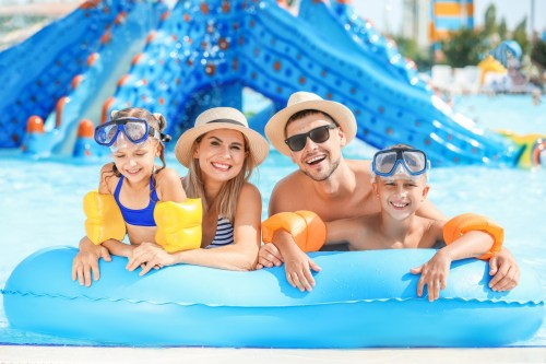 A happy family of four in swim gear smiles while floating on a blue inflatable in a water park.