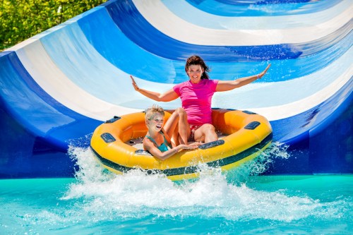 A woman and a child joyfully ride a yellow inflatable raft down a colorful water slide, splashing into the pool below.